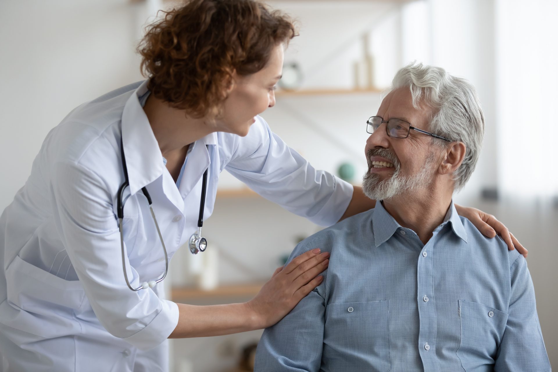 Smiling young nurse caregiver supporting mature patient during homecare visit, friendly woman doctor wearing white uniform with stethoscope touching senior man shoulders, psychological help